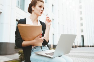 Thoughtful Female Student Sits In Park Near University And Prepares For Exams. Young Teacher Prepares For Lecture And Searches For Information On Internet With A Laptop Computer Sitting On Urban Street